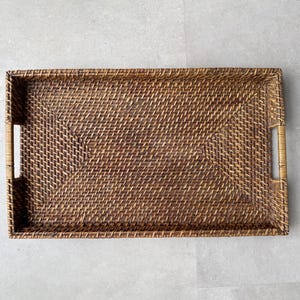 May include: A rectangular woven tray crafted from dark brown rattan material. The tray features a tight, diagonal weave pattern across the base and sides, with integrated cutout handles on the two shorter ends for easy carrying and handling.