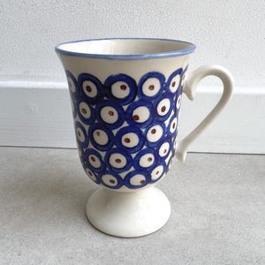 Könnte beinhalten: Eine weiße Keramiktasse mit blauem und braunem Muster. Die Tasse hat einen Sockel und einen gebogenen Henkel. Das Design zeigt blaue Kreise mit braunen Zentren. Der Rand und der Griff sind weiß mit blauem Rand.