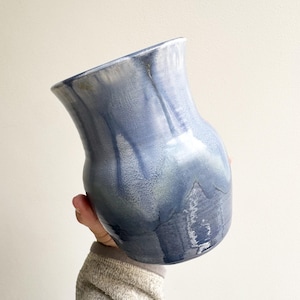 Op de afbeelding: Een blauwe keramische vaas met een uitlopende rand en een afgeronde body. De vaas heeft een glanzende afwerking met strepen van lichtblauw en wit, waardoor een gemarmerd effect ontstaat. De vaas wordt door een persoon vastgehouden.