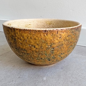 Op de afbeelding: Een handgemaakte, getextureerde keramische kom met een brede, afgeronde vorm. De kom heeft een gevlekte, aardse kleurenpalet van goudbruin, beige en vleugjes groen. De binnenkant heeft een lichtere, gespikkelde uitstraling.