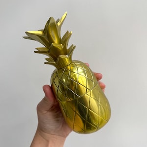 Op de afbeelding: Een metallic goudkleurig object in de vorm van een ananas met een textuur van ruitpatronen op het lichaam en een puntige, bladvormige bovenkant. Het oppervlak heeft een gladde, reflecterende afwerking die het licht opvangt en accentueert.