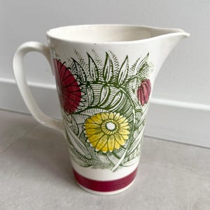 May include: A white ceramic pitcher with a curved handle and spout. The pitcher is decorated with red, yellow, and green floral designs. A red band accents the base. This decorative pitcher is ideal for serving beverages.