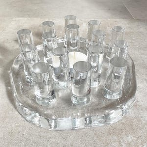 May include: A clear glass candle holder with 12 cylindrical glass holders arranged in a circle around a central candle. The base is oval-shaped and made of clear glass. The candle holder is on a light gray surface.