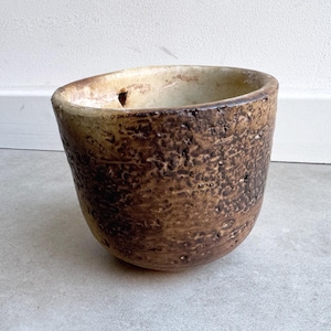 Op de afbeelding: Een ronde, komvormige keramische pot met een dikke, onregelmatige rand. De buitenkant heeft een zwaar getextureerd, donkerbruin en beige gevlekt oppervlak met een ruwe afwerking. De binnenkant is glad, lichtbeige en vertoont een kleine donkere vlek.