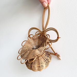 Könnte beinhalten: Ein geflochtener Korb aus Rattan mit dekorativem Schleifenhenkel. Der Korb hat einen runden Boden und einen gewellten oberen Rand, mit einem kleinen silbernen Gegenstand im Inneren. Der Korb wird von einer Hand vor weißem Hintergrund gehalten.