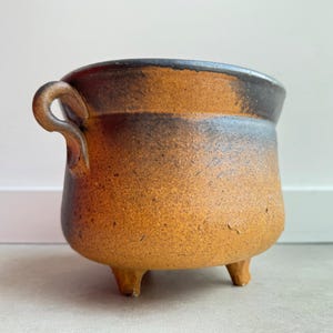 Può includere: Un vaso in ceramica marrone e nero con un'unica maniglia e tre gambe. Il vaso ha una smaltatura screziata.