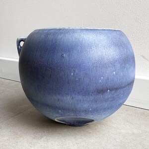 May include: A round, blue ceramic vase with a handle. The vase has a smooth, glazed finish with subtle color variations. The overall shape is spherical, with a slightly wider top. The vase is suitable for home decor.