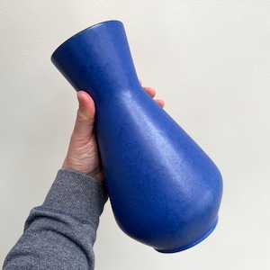May include: A cobalt blue vase with a textured surface. The vase has a flared top and a rounded base. The vase is held up against a white background. The vase is approximately 10 inches tall.
