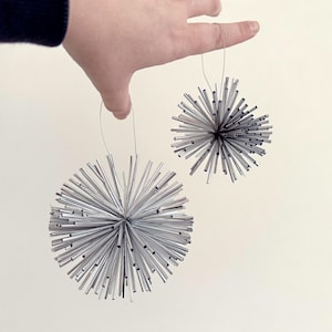 Pode incluir: Dois ornamentos esféricos prateados com um design espinhoso em forma de estrela. Cada ornamento é feito de numerosas varetas metálicas finas que irradiam de um ponto central. São suspensos por fios finos.