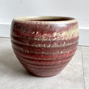 Könnte beinhalten: Handgefertigte Keramikschale mit breiter, runder Form. Die Schale hat horizontale Bänder in tiefroten und beigen Farben, die ein strukturiertes, erdiges Aussehen erzeugen. Der Innenraum ist hellbeige.