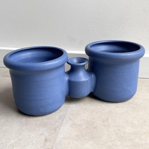 May include: A blue ceramic planter with three connected sections. Two larger, round openings are connected by a smaller, vase-like section. The planter has a matte finish and is suitable for indoor plants.