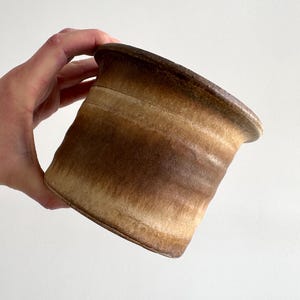 Könnte beinhalten: Ein handgefertigter Keramik-Pflanztopf mit einer einzigartigen, ausgestellten Form. Die Außenseite weist einen Farbverlauf von Brauntönen auf, von hellem Beige bis Dunkelbraun, wodurch eine natürliche, erdige Ästhetik entsteht. Der Topf wird von einer Hand vor weißem Hintergrund gehalten.