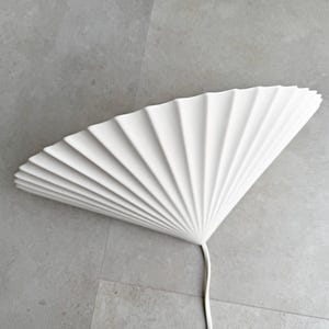Op de afbeelding: Een witte, waaiervormige wandlamp met een geplooid ontwerp. De lamp is gemaakt van een stevig materiaal en heeft een gladde, matte afwerking. Een wit snoer loopt vanaf de basis van de lamp. De lamp is op een grijze muur gemonteerd.