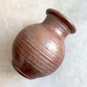 May include: A small, handcrafted ceramic vase with a rounded body and a slightly flared rim. The vase has a speckled, reddish-brown glaze with horizontal striations, giving it a rustic appearance. The vase is approximately 5 inches tall.