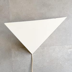 Pode incluir: Uma luminária de parede triangular com um acabamento liso e mate em branco sujo. A peça apresenta uma superfície larga e plana que se estreita para uma ponta na parte inferior, onde um cabo de alimentação fino e claro sai do centro da base.