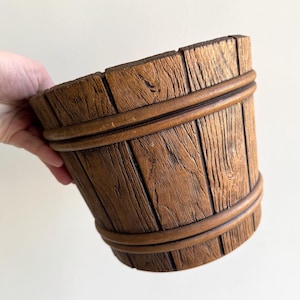 May include: A wooden bucket with a rustic design. The bucket is made of vertical wooden planks held together by two encircling bands. The wood has a warm brown color and visible grain, giving it a vintage appearance. The bucket is held by a hand.