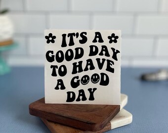 May include: A square, off-white sign with the phrase "IT'S A GOOD DAY TO HAVE A GOOD DAY" in black, retro-style lettering. The sign is decorated with flower accents and a smiley face. It rests on a stack of two wooden coasters.