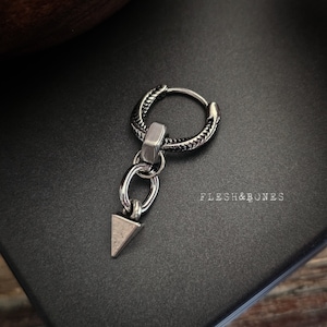 Könnte beinhalten: Silberner Creolenohrring mit strukturiertem Design und einem baumelnden Dreiecksanhänger. Der Ohrring liegt auf einer schwarzen Oberfläche mit dem aufgedruckten Text "FLESH&BONES".