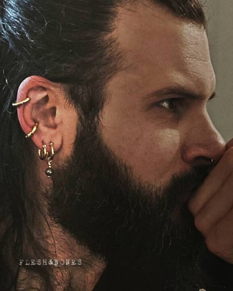 May include: A man with long black hair and a beard is wearing multiple gold hoop earrings in his left ear. The earrings are stacked on top of each other and include a small gold bead dangling from the bottom hoop. The man is looking down and to the right, and his hand is covering his mouth.