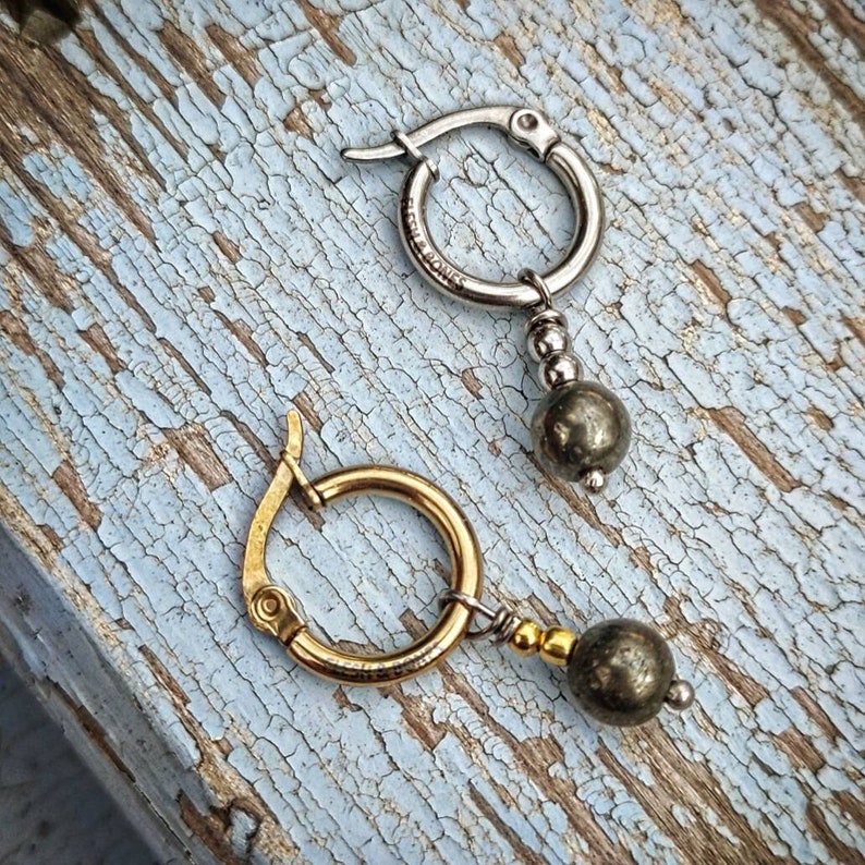 May include: A pair of hoop earrings with a dangling pyrite stone. The earrings are made of silver and gold metal. The pyrite stones are a dark gray color.