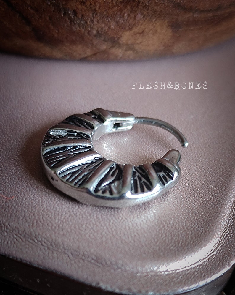 May include: Silver hoop earring with a textured, geometric design. The earring is made of stainless steel and has a hinged closure.