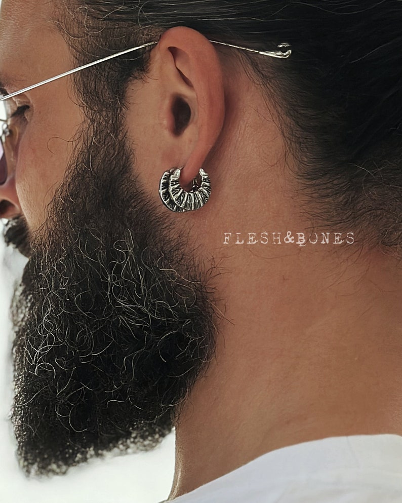 May include: Silver hoop earrings with a textured, ridged design. The earrings are worn in the ear of a person with a beard.