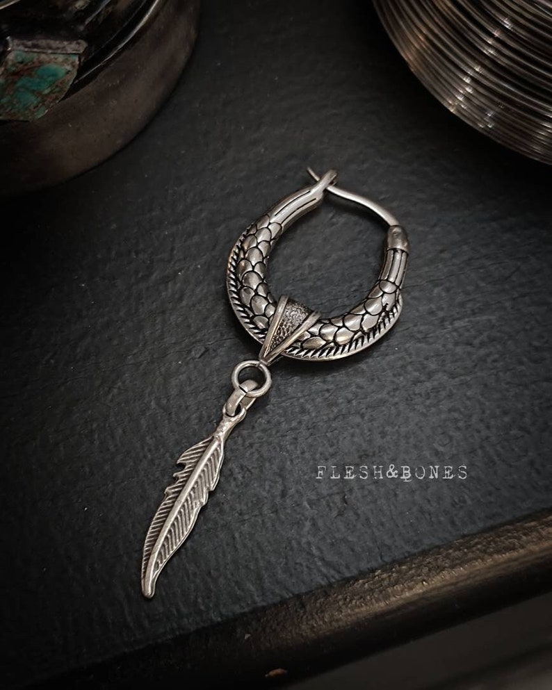 Puede incluir: Pendiente de aro de plata con un dise&ntilde;o texturizado y un colgante de pluma. El pendiente est&aacute; sobre una superficie oscura con el texto "Flesh & Bones" visible.