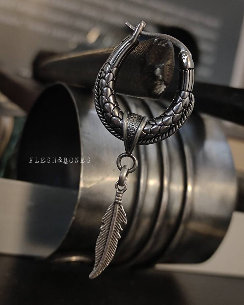 Puede incluir: Un aro de plata para la oreja con un dise&ntilde;o texturizado y un colgante de pluma. El aro est&aacute; hecho de metal y tiene un acabado oxidado oscuro.