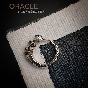 Op de afbeelding: Zilveren oorring met gouden accenten en ingewikkelde details. De oorring is afgebeeld op een zwart-wit gestreept oppervlak. De tekst "ORACLE FLESH&BONES" is zichtbaar.