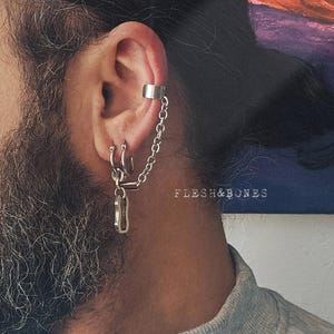 Puede incluir: Un pendiente de aro de plata con una cadena unida a un pequeño dije en forma de llave. El aro se lleva en la parte superior de la oreja, y la cadena cuelga hasta el lóbulo inferior. El pendiente de aro es un estilo de joyería popular para hombres y mujeres.