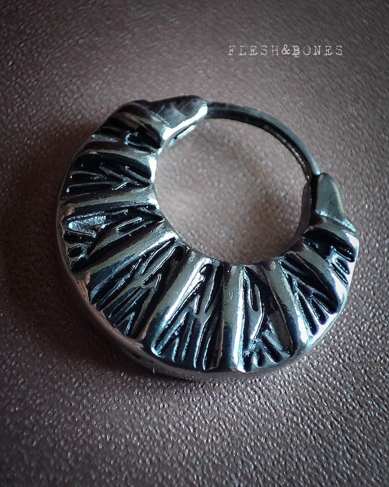 May include: Silver hoop earring with a textured, geometric design. The earring is a simple circle with a raised pattern.