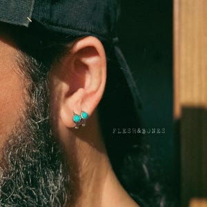 Puede incluir: Una persona lleva una gorra de béisbol negra con dos pendientes de piedra turquesa en la oreja izquierda. El texto "FLESH&BONES" es visible en la imagen.