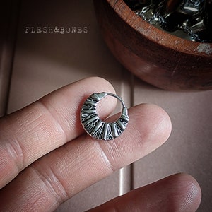 May include: Silver hoop earring with a textured design. The earring is on a finger.