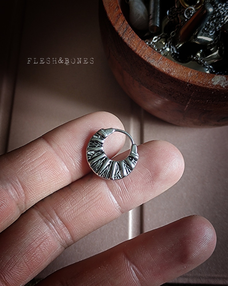 May include: Silver hoop earring with a textured design. The earring is on a finger.