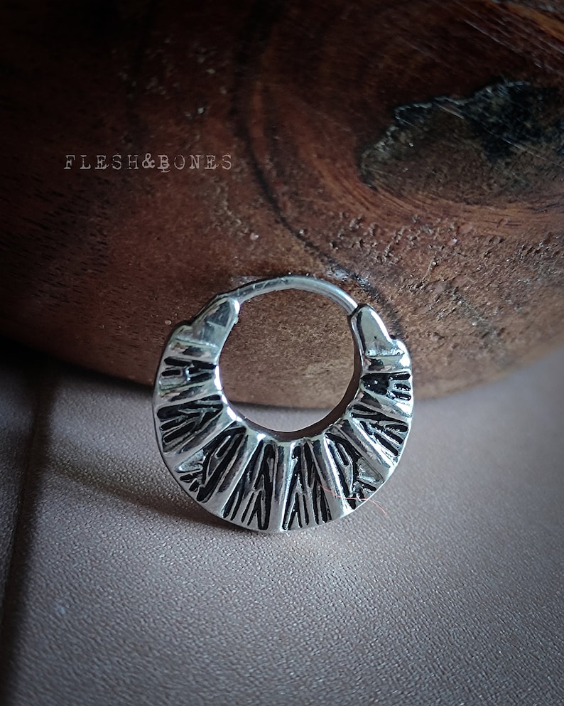 May include: Silver hoop earring with a textured, geometric design. The earring is circular and has a slightly curved shape.