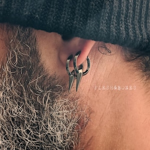 Può includere: Primo piano di un orecchio con tre orecchini a cerchio neri con un design a punta. Gli orecchini sono in metallo scuro. Il testo "FLESH&BONES" è visibile sullo sfondo.