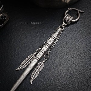 Può includere: Orecchino in argento con un lungo pendente sottile in argento che comprende una catena, una piccola perla in argento e due piume in argento. Il pendente ha la forma di una lancia o di una punta di freccia. L'orecchino è contrassegnato da "FLESH & BONES".