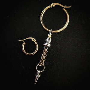 May include: A pair of gold hoop earrings, one with a dangling charm featuring a silver and gold chain, a silver and gold bead, and a silver spike. The hoop earring with the charm has the words "Flesh & Bones" engraved on the hoop.