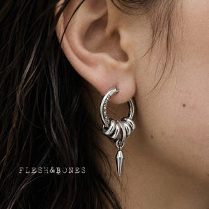Op de afbeelding: Een zilveren oorring met de gegraveerde tekst FLESH & BONES op de band. De ring is versierd met vier kleinere zilveren ringetjes en een centrale zilveren hanger in de vorm van een punt die onderaan hangt. Het metaal heeft een gepolijste afwerking.