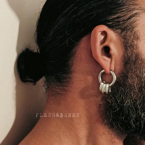 May include: Silver hoop earring with three stacked rings. The earring is worn on the ear of a person with dark hair pulled back and a beard. The text "FLESH&BONES" is printed in the image.