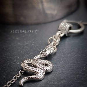 Puede incluir: Collar de serpiente de plata con cadena y cierre decorativo. La serpiente est&aacute; enroscada y tiene una superficie texturizada. El collar est&aacute; sobre un fondo gris oscuro.