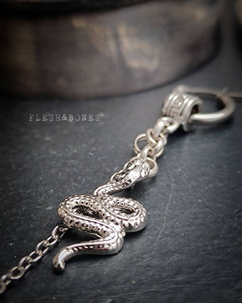 Puede incluir: Collar de serpiente de plata con cadena y cierre decorativo. La serpiente est&aacute; enroscada y tiene una superficie texturizada. El collar est&aacute; sobre un fondo gris oscuro.