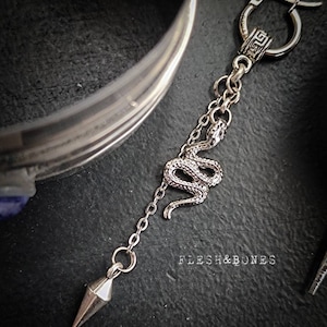 Puede incluir: Pendiente de aro de plata con un dije de serpiente y un colgante puntiagudo. El pendiente tiene un dise&ntilde;o de llave griega en el aro. El texto "Flesh & Bones" es visible en el fondo.