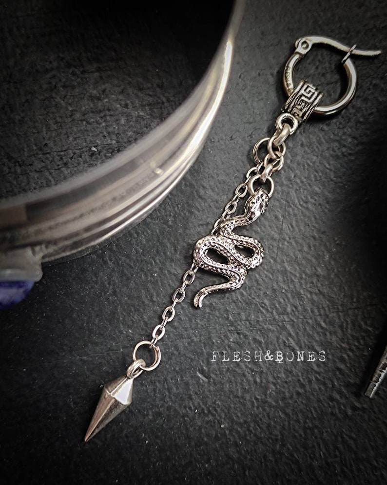 Puede incluir: Pendiente de aro de plata con un dije de serpiente y un colgante puntiagudo. El pendiente tiene un dise&ntilde;o de llave griega en el aro. El texto "Flesh & Bones" es visible en el fondo.