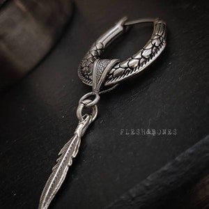 Puede incluir: Pendiente de aro de plata con un dise&ntilde;o texturizado de escamas y un colgante de pluma. El pendiente est&aacute; sobre un fondo oscuro con el texto "Flesh & Bones" visible.