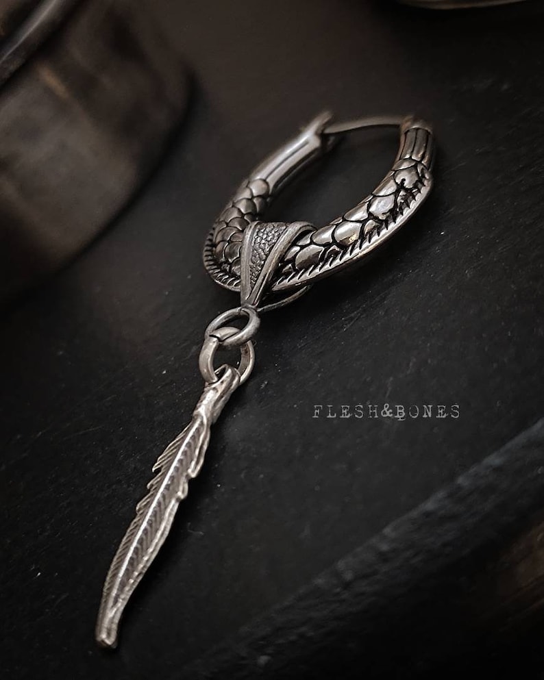 Puede incluir: Pendiente de aro de plata con un dise&ntilde;o texturizado de escamas y un colgante de pluma. El pendiente est&aacute; sobre un fondo oscuro con el texto "Flesh & Bones" visible.