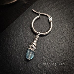 Op de afbeelding: Zilveren oorring met een hangende blauwe edelsteenbedel. De bedel heeft een zilveren kraal en een decoratieve zilveren dop.