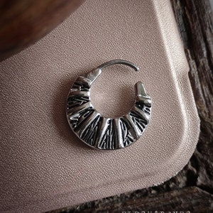 May include: Silver hoop earring with a textured, geometric design. The hoop is slightly curved and has a hinged closure.