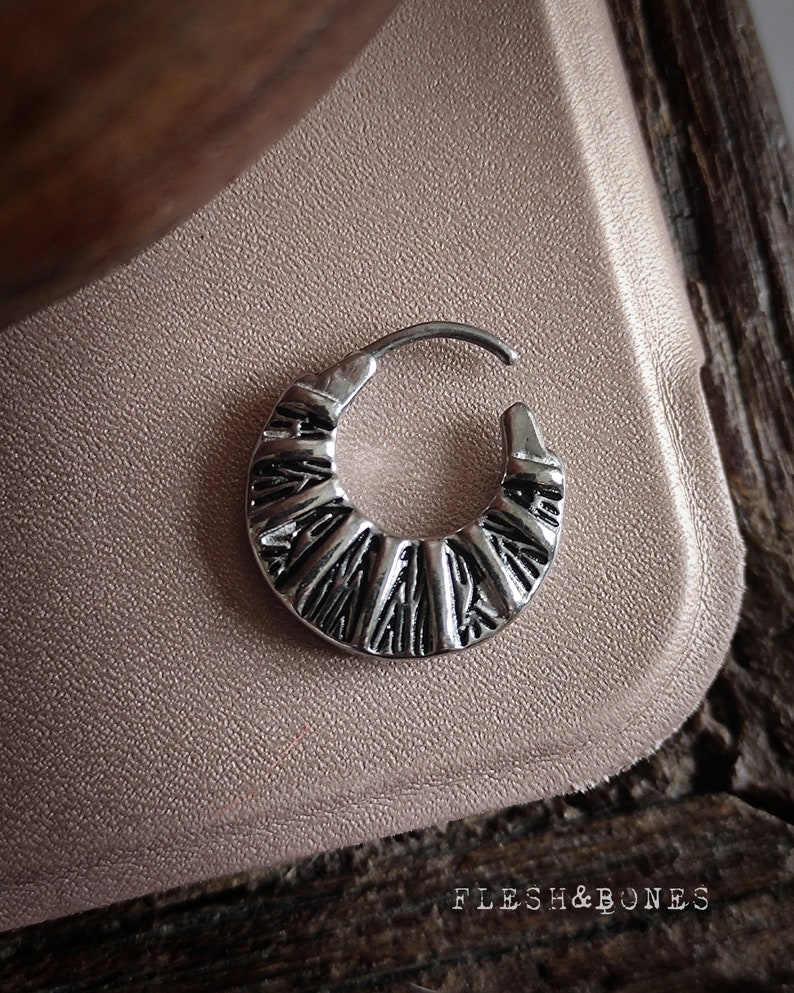 May include: Silver hoop earring with a textured, geometric design. The hoop is slightly curved and has a hinged closure.
