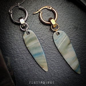 Könnte beinhalten: Ein Paar Ohrringe mit einem silbernen und goldenen Kreis und einem langen, tropfenförmigen Anhänger mit einem blauen und weißen Marmormuster. Die Ohrringe liegen auf einer dunkelgrauen Oberfläche.
